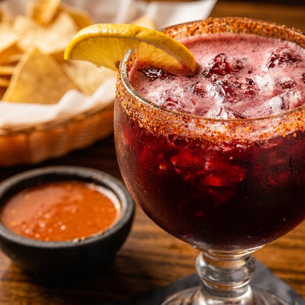 A red margarita with a chili-lime rim and lemon wedge, next to tortilla chips and salsa.