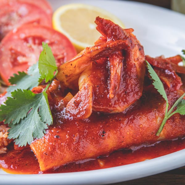 Enchiladas covered in red sauce with Mexican rice, cucumber, tomato, lemon, and cilantro on a white plate.