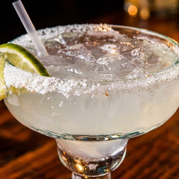 Close-up of a margarita with a salted rim, lime wedge, and straw on a dark wooden table.
