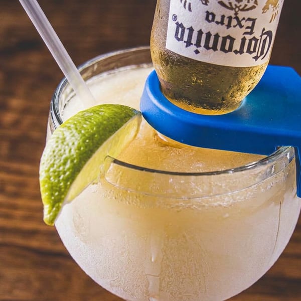 Frozen margarita with an inverted Coronita beer bottle, straw, and lime wedge on a wooden table.