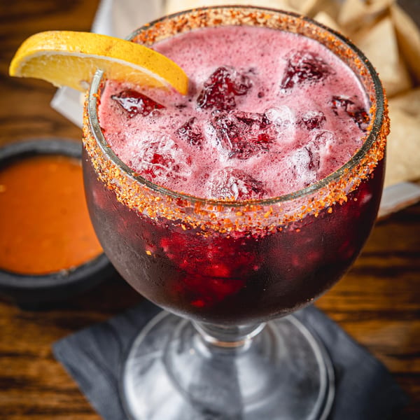 Red berry cocktail with ice, an orange wedge, and a chili-salt rim served in a stemmed glass on a wooden table.