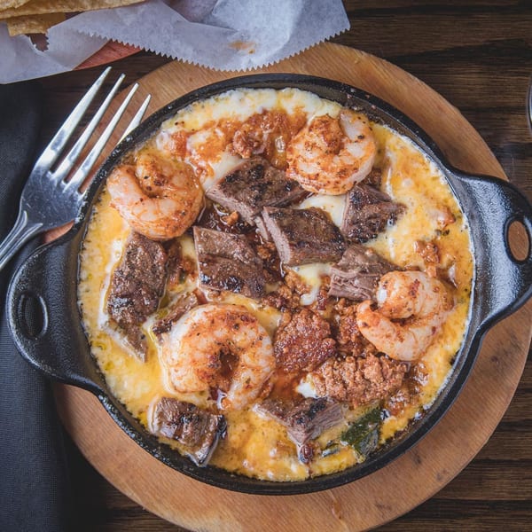 Overhead view of grilled shrimp and steak pieces in creamy sauce, served in a cast-iron skillet with tortillas and utensils.