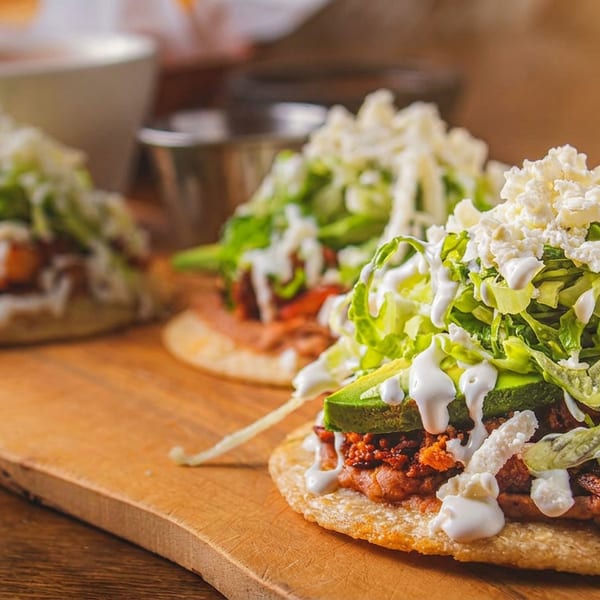 Three Mexican sopes topped with meat, lettuce, avocado, sour cream, and cheese on a wooden board.