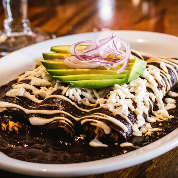 Black mole enchiladas topped with cream, crumbled cheese, avocado slices, and red onion on a white plate.