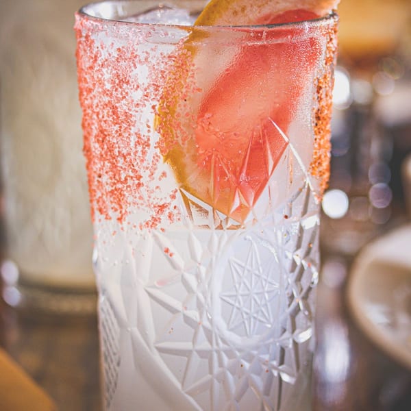 Pink citrus cocktail in a patterned glass with a sugar-coated rim, grapefruit garnish, and straw on a dimly lit bar.