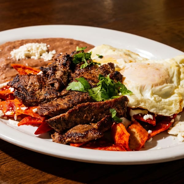 A white plate with steak, fried eggs, refried beans, and red chilaquiles on a wooden table.