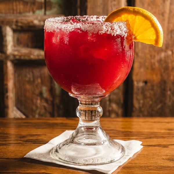 A vibrant red margarita with a salted rim and orange slice sits on a wooden table.